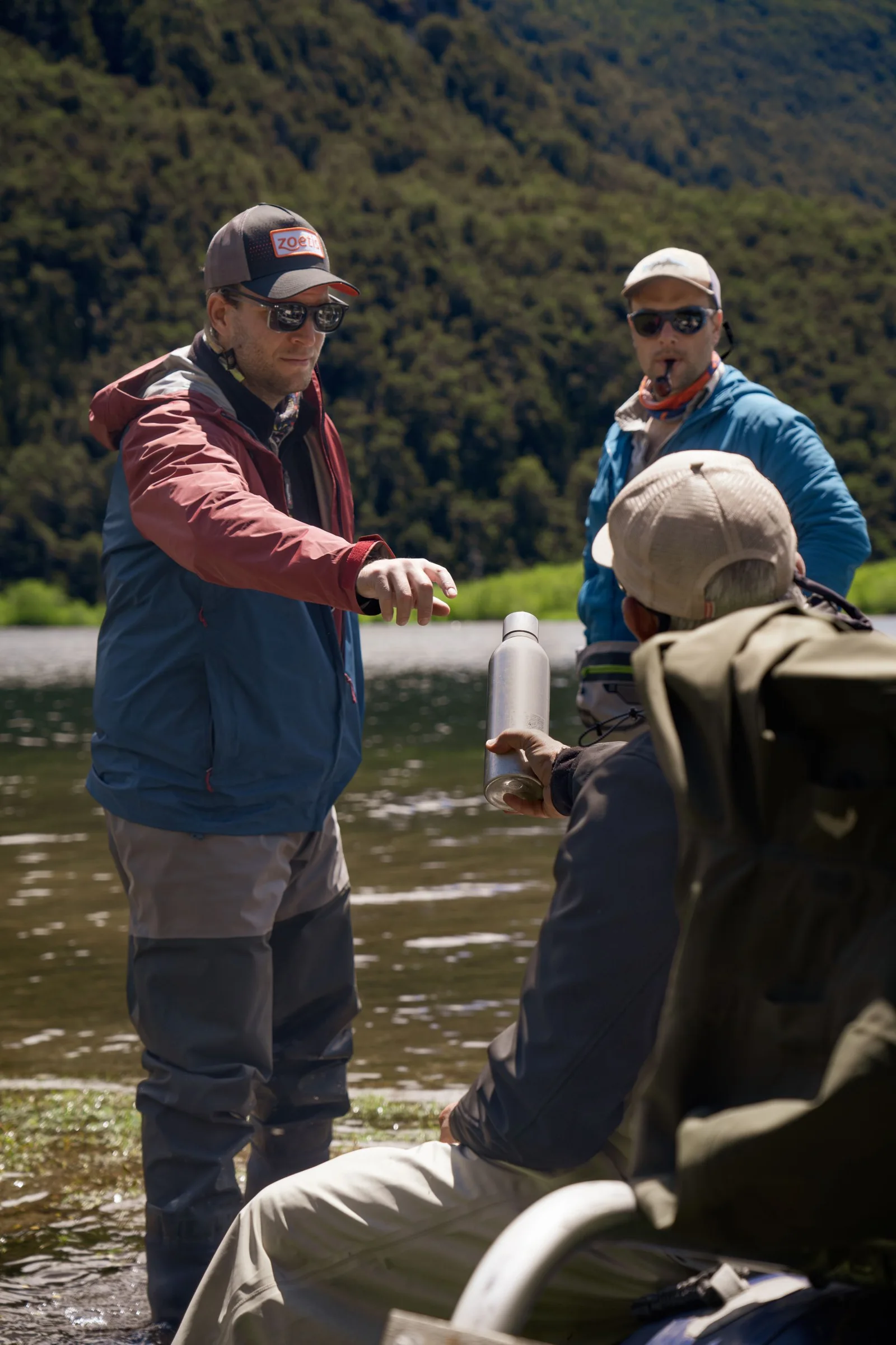 Three anglers fly fishing beside a river beneath green mountains.
