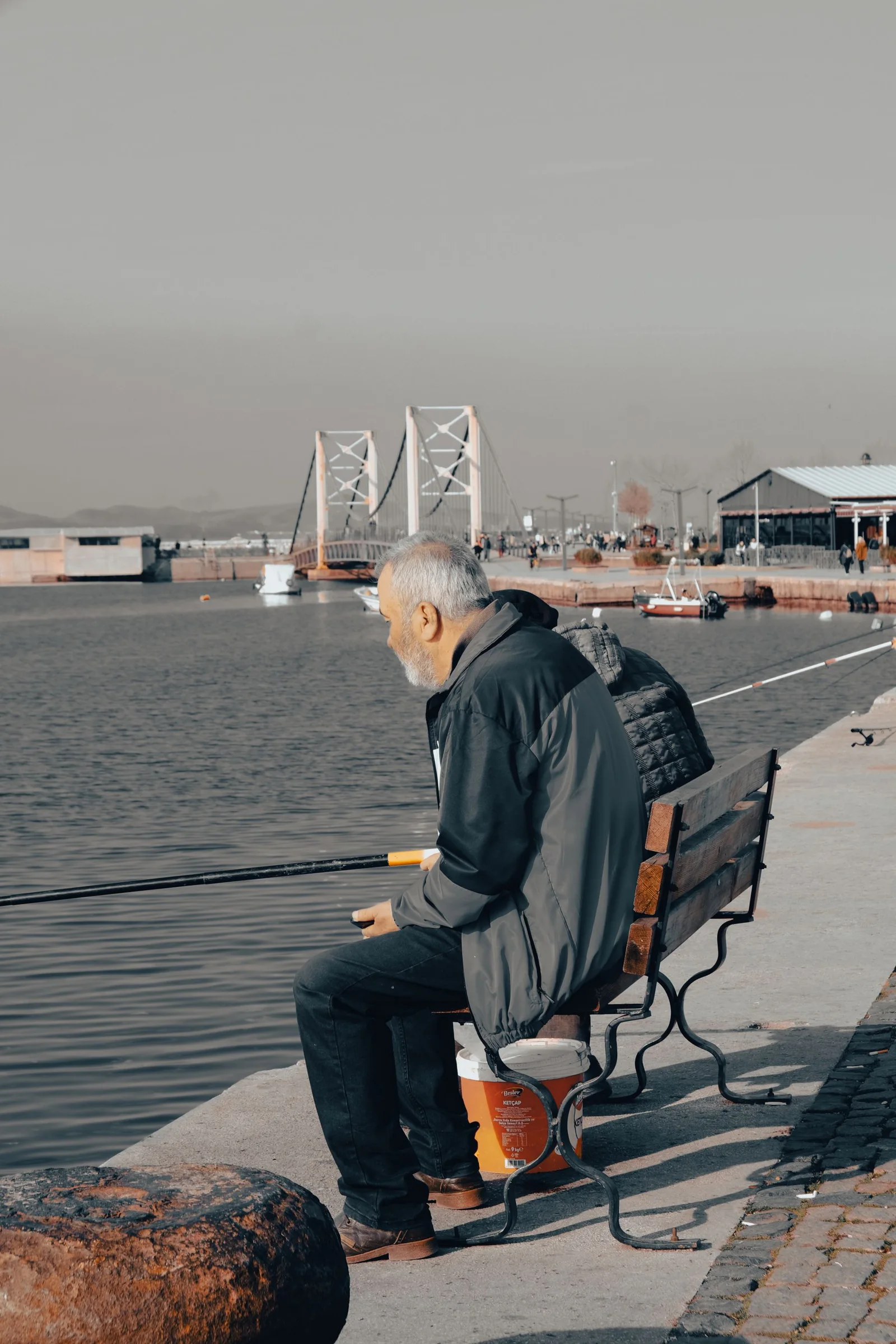 An older angler sitting by a riverbank with a bridge in the background.