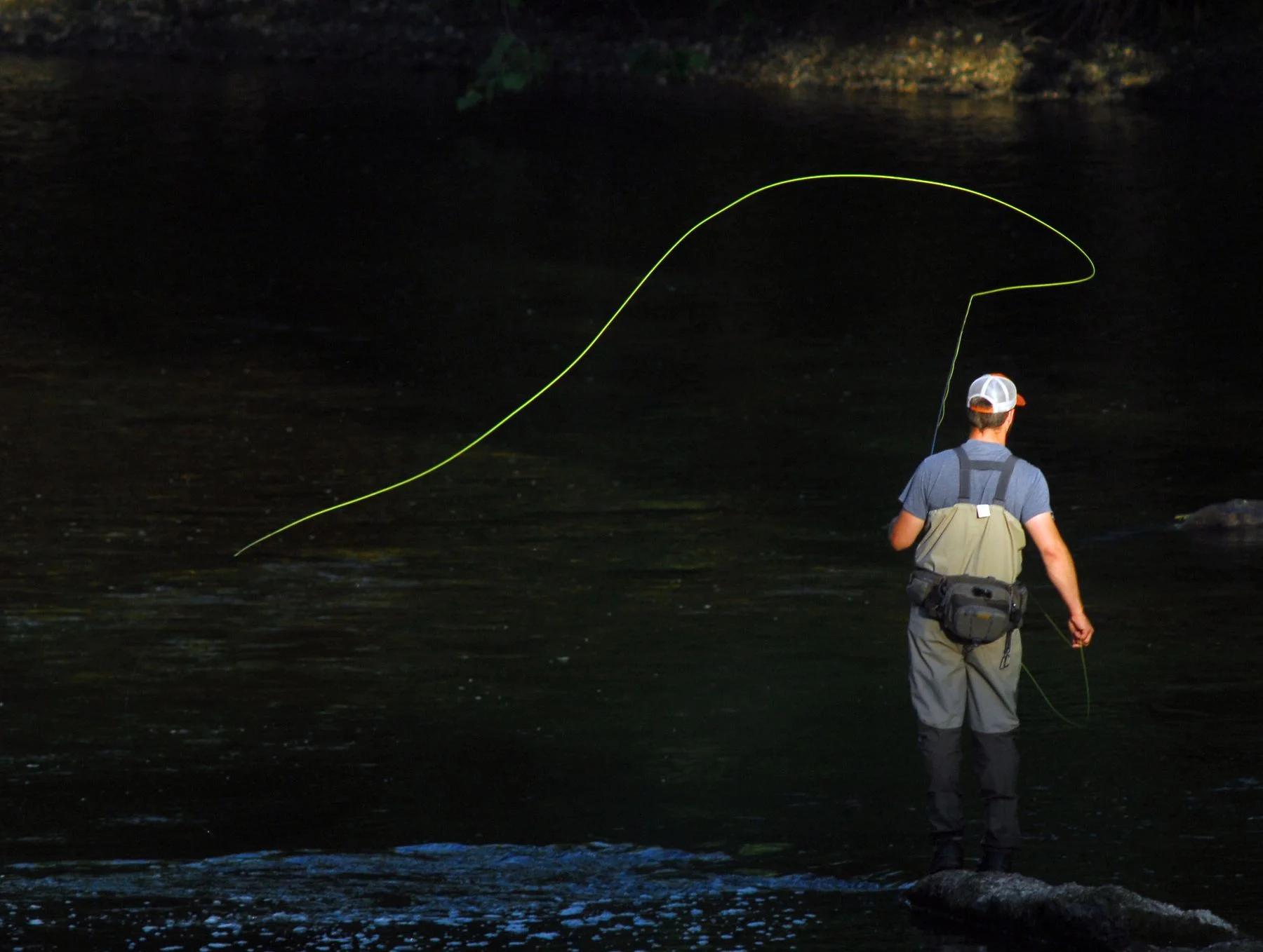 An angler in waders casting a fishing line in a river.