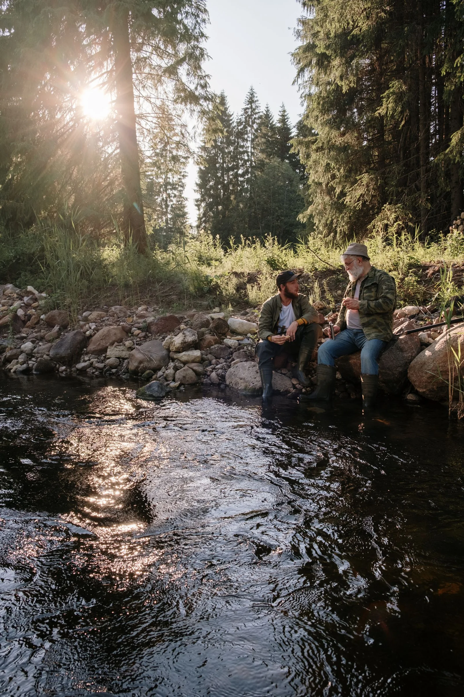 Two people sitting with fishing gear beside a wooded river.