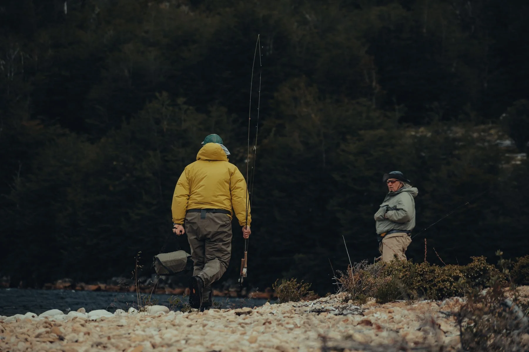 Two anglers carrying fishing rods beside a wooded riverbank.