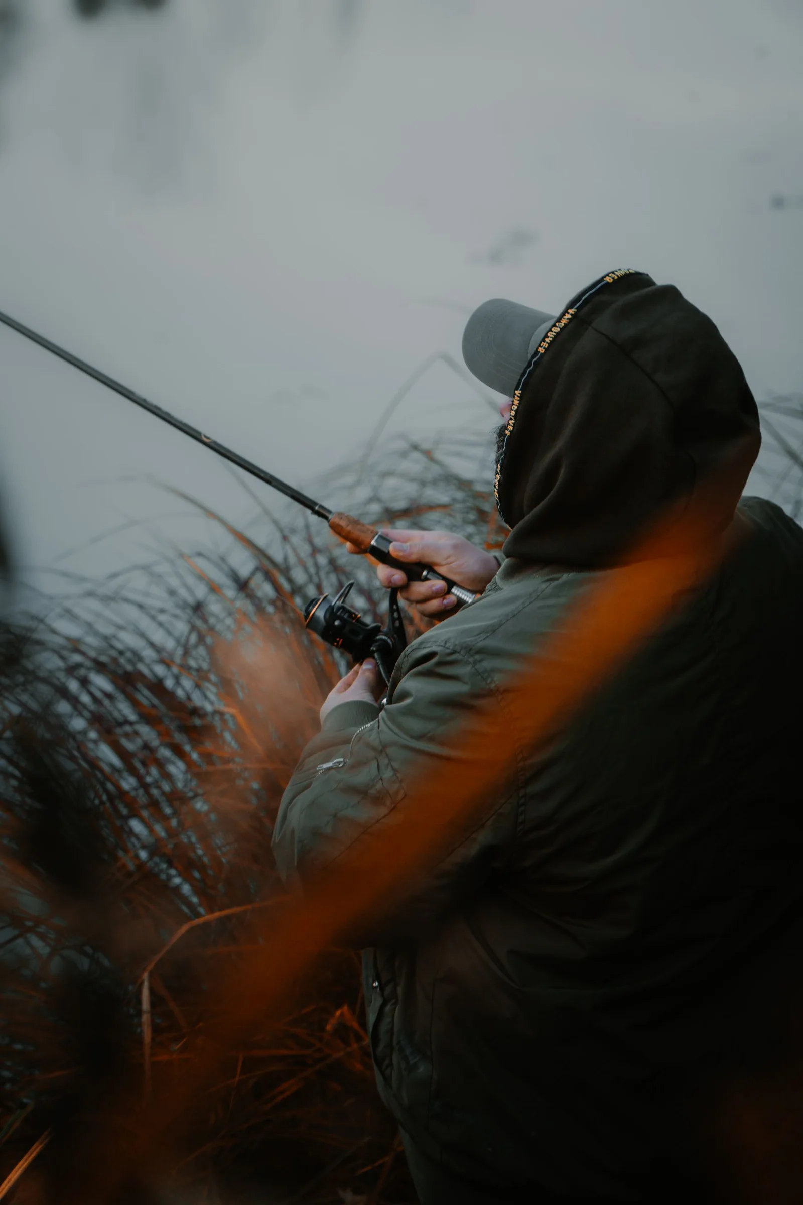 An angler in a hooded jacket fishing beside still water and autumn foliage.
